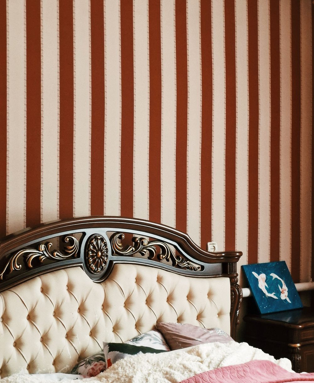 Bedroom with rust-and-cream striped wallpaper hung behind an ornately carved, tufted headboard