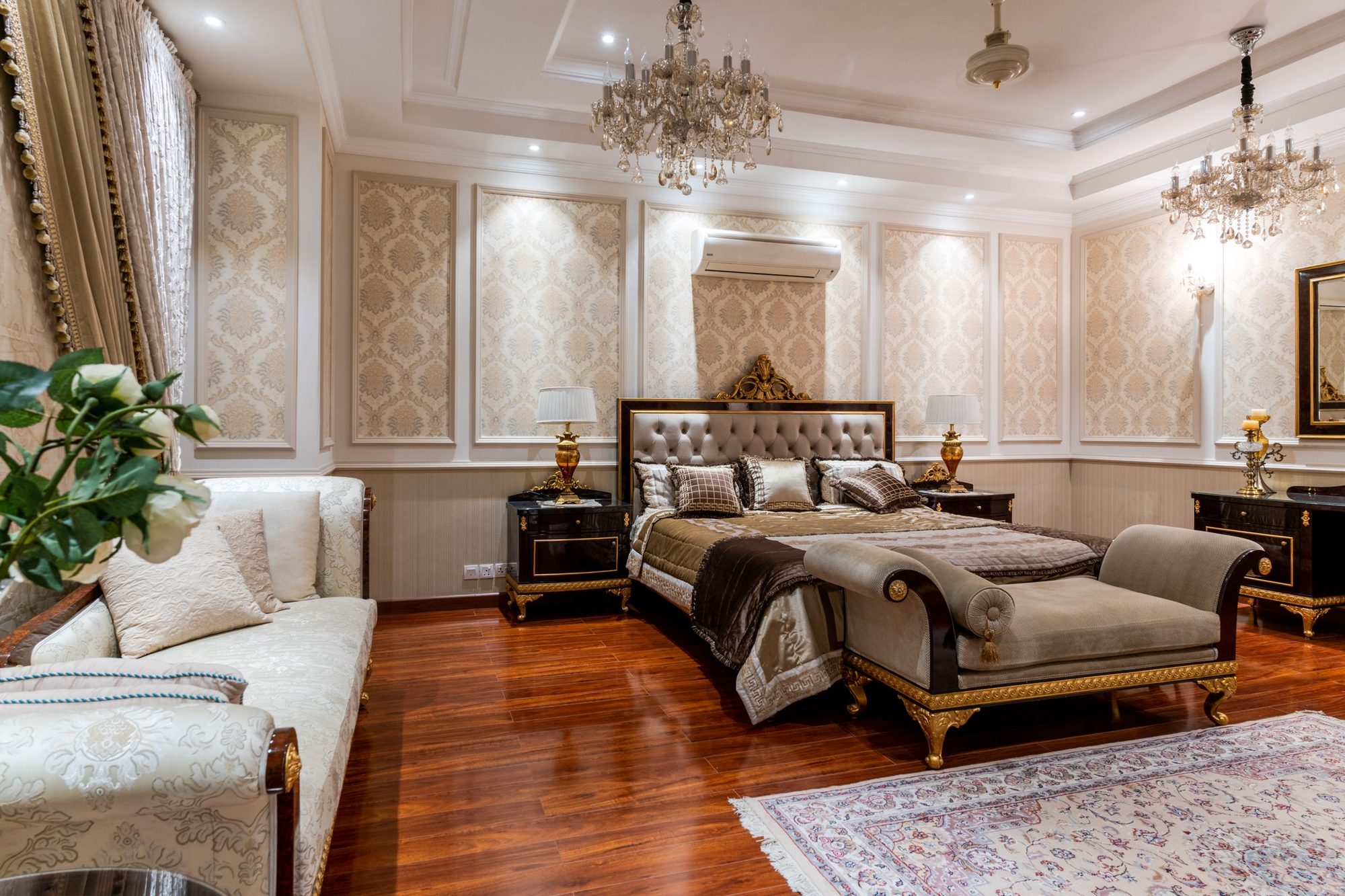 Elegant residential bedroom with cream damask wallpaper panels framed by white molding, a tufted headboard and crystal chandeliers